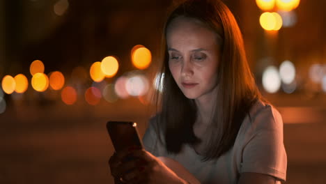 A-young-woman-looks-into-the-smartphone-and-writes-text-messages-on-the-Internet-against-the-backdrop-of-the-night-city.-Girl-businessman-working-on-vacation-remote-work-via-mobile-phone.-Gadget-addiction